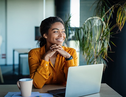 Woman wearing golden shirt smiling looking out window at work desk